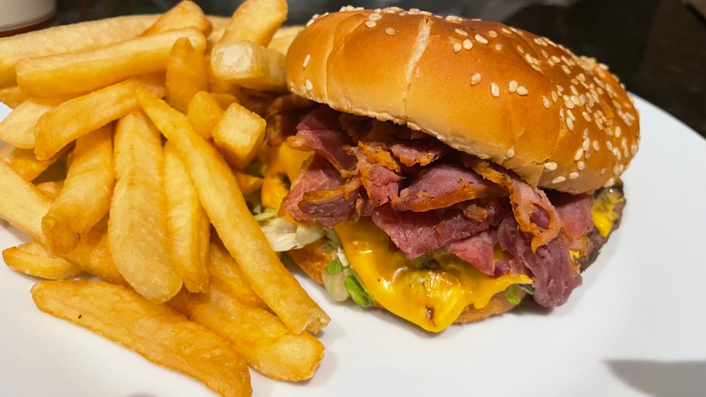 A crown burger overflows with meat and cheese and sits beside a handful of fries atop a white plate.